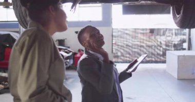 Servis danışmanı tableti tıkırdatıyor ve teknik ekip asansörü tamir ederken arabanın altını işaret ediyor. Otomotiv, takım çalışması, profesyonel, endüstriyel, bakım, işbirliği, denetleme