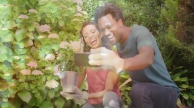 Arka bahçede diz çöküp selfie çekmek için akıllı telefon kullanan ve sosyal medya için çiçek açan bir çift. Bahçıvanlık, açık hava, doğa, mutluluk, arkadaşlık, yaşam tarzı, canlı