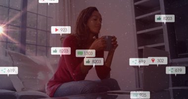 Gri kupa tutan ve sosyal medya simgeleriyle oturma odasında gümüş dizüstü bilgisayarı kontrol eden bir kadın. Rahat, modern, teknolojik, gündelik, rahatlatıcı, iletişim, çalışma