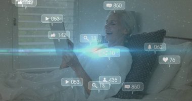 Yatakta tableti tutan yaşlı bir kadın, gri örülmüş yastıkla, yüzen dijital kaplamalarla. Rahatlama, teknoloji, dijital, konfor, yaşam tarzı, bağlantı, modern