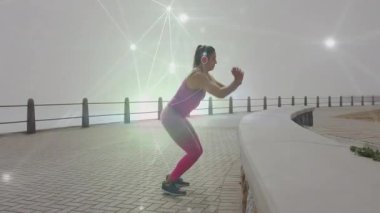 Kadın, deniz kenarındaki spor salonunda öne eğilip kulaklıklarla kıvrımlı duvara zıplıyor. Egzersiz, fitness, aktif giysi, şehir, açık hava, motivasyon, dinamik