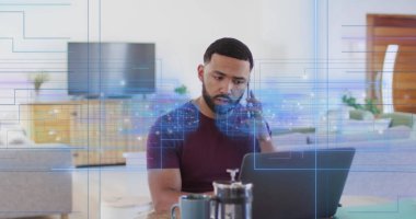 Akıllı telefondan konuşan kestane rengi tişörtlü adam evdeki masada dijital kaplamalarla dizüstü bilgisayar kullanıyor. Teknoloji, uzaktan çalışma, modern, yaşam tarzı, dijital, üretkenlik, gündelik