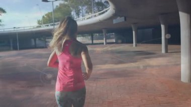 Üst geçidin altındaki terracotta plazasında koşan bir kadın, formun etrafında titreşen sağlık düğümlerini başlatıyor. Fitness, urban, motion, technology, connectivity, wellness, activewear