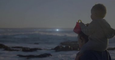 Anne omuzlu bebek kırmızı kovayı kaldırıyor kayalık sahil yaşam tarzının üzerinden güneşe uzanıyor. Sahil, şafak, siluet, dinginlik, bağ, macera, ufuk