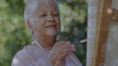 Kıdemli kadın fırçasını palete daldırıyor ve bokeh 'in arka bahçesinde tuvale manzara çiziyor. Yaratıcı, sakin, hobi, izlenimci, açık hava, pastel, boş zaman