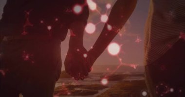 Gün batımı kıyı şeridinde el ele tutuşan çift teknolojide kırmızı moleküler ışıkları çalıştırıyor. Bağlantı, samimiyet, uyum, dinginlik, teknoloji, sıcaklık, romantizm