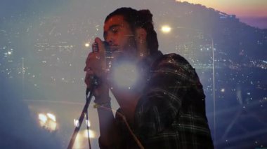 Erkek şarkıcı mikrofon standında mırıldanıyor, şehir ışıkları siluet etrafında dönerken vokalleri yoğunlaştırıyor. Gece hayatı, şehir, performans, müzik, vokal, sahne, şehir