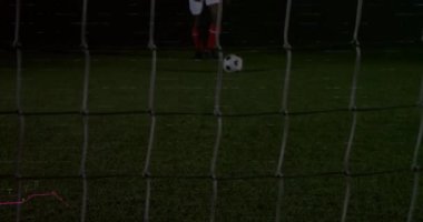 Top ışıkların altında koşuyor ve kaleci dalarken kale üzerinden gol atmak için topa vuruyor. Atletizm, rekabet, kararlılık, stadyum, aksiyon, spor, yoğunluk