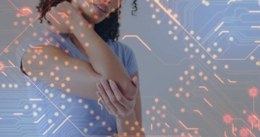 Standing woman wearing blue shirt holding right elbow inside room, with digital circuit overlay. Technology, innovation, digital, healthcare, modern, circuit, connectivity
