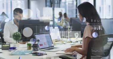 Typing woman in blush blouse analyzing report on laptop in office, with notebook, monitors, plant. Collaboration, professionalism, modernity, teamwork, analytics, workspace, productivity