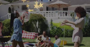 Erkekler gitar çalarken, iki kadın çimenlikte şişme top geçirip yaşam tarzı içeriği yaratıyor. Açık hava, boş zaman, neşeli, rahat, canlı, dostluk, yaz