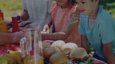 Çocuklar piknik yemekleri için uzanırken veriler üst üste binerken erkekler sağlık kavunu servis ediyor. Aile, piknik, açık hava, boş zaman, güneş ışığı, toplanma, sağlıklı