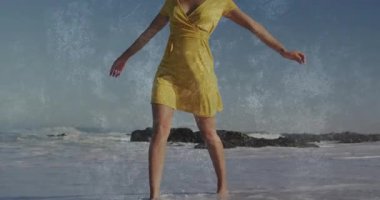 Bacağını dalgalara doğru kaldıran bir kadın, su üstü örtülü, yürüyen bir sahile sarı bir elbise giydirmiş. Deniz manzarası, zarafet, akıcılık, esinti, dinginlik, açık hava, yaşam tarzı.