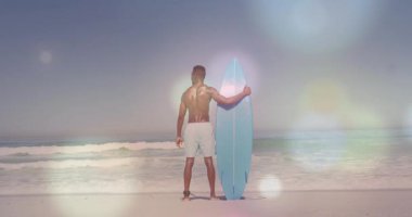 Sahilde sörf tahtası tutan tişörtsüz bir erkek sörfçü güneş ışığı gibi ufuk çizgisine bakıyor. Bu da lenslerin parlamasına neden oluyor. Sahil, sahil, okyanus, dalgalar, kıyı şeridi, güneş ışığı, macera