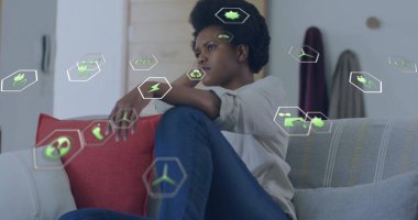 Oturan kadın evdeki gri kanepeye dikkatlice bakıyor, kırmızı yastıkla, yüzen eko ikonlarıyla. Çevre, sürdürülebilirlik, farkındalık, şehir, minimalizm, enerji, düşünceli