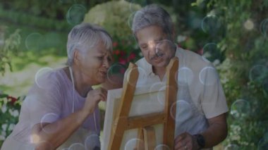 Bahçe sehpasında yelken baloncuklu sanat eserleri için fırça kapıp tuval boyayan yaşlı çift. Zenith, sükunet, yaratıcılık, arkadaşlık, açık hava, empresyonist, ayyaş