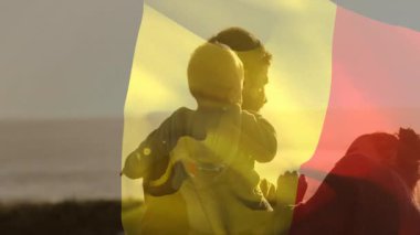 Bayrak dalgalanması, baba beşikte bebeği tetiklerken anne de okyanus manzarasına sağlık için rehberlik ediyor. Aile, ebeveynlik, bağlılık, açık hava, gün batımı, dinginlik, şefkat