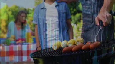 Mısırları ve sosisleri ters çeviren maşa kullanan bir adam aynı zamanda çocuklara anne tabaklarıyla ızgara yapmayı öğretiyor. Aile, açık hava, boş zaman, ızgara, birliktelik, yaz, kırsal