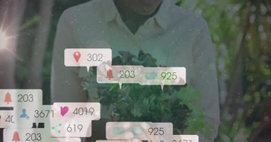 Bahçedeki yapraklı yeşillikleri indiren bir kadın sosyal medya için lokasyon çanı ve kamera ikonlarını ateşliyor. Organik, sağlık, yaşam tarzı, doğa, açık hava, içerik oluşturma, bağlılık