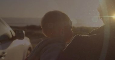 Anne deniz kıyısında arabadan iniyor, bebeği kucaklıyor, gün batımını merceklerle seyrediyor sağlık için. Aile, bağ, açık hava, dinginlik, manzara, siluet, ebeveynlik