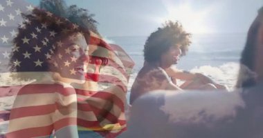 Sahil havlularında oturmuş muhabbet eden arkadaşlar ABD bayrağı dalgaların üzerinde dalgalanmaya başlarken gülüyorlar. Gündelik, açık hava, yaşam tarzı, hayat dolu, rahat, sosyal, yaz