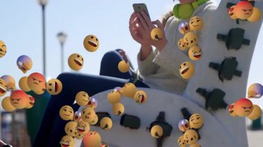 Akıllı telefonu kaydıran ve sosyal medya tanıtımı için bankın etrafında hareketli reaksiyon sembolleri yaratan bir kız. Teknoloji, gençlik, iletişim, ifade, rahatlama, açık hava, canlı