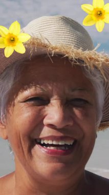 Composite video of flowers icons falling against african american senior woman smiling at the beach. travel and vacation concept