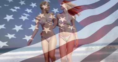 Plajda yürüyen iki kadın, şeffaf Amerikan bayrağı altında kareye girerek seyahati hızlandırıyor. Arkadaşlık, sahil, sahil, tatil, boş zaman, güneşlenme, rahatlama.