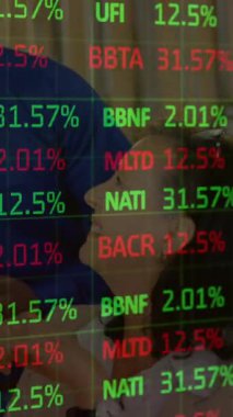 Bir grup işletme yöneticisi, borsa verileriyle çevrili bir şekilde ofiste eğleniyor. Veriler yuvarlanıyor ve işliyor, kırmızı ve yeşil renkler pazarın farklı yönlerini temsil ediyor.