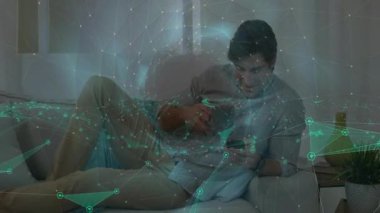 Kanepede oturan yetişkin bir adam telefonu kaydırıyor ve teknoloji bağlamında ağ kaplamalı bardaktan içiyor. Bağlı, gevşeme, modern, dijital, iletişim, gündelik, boş zaman