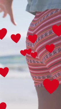 Sahilde el ele tutuşan Afrikalı Amerikalı çiftin orta kesiminde kırmızı kalplerin animasyonu. Aşk, birliktelik, romantizm ve tatil konsepti dijital olarak oluşturulmuş video.