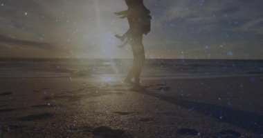 Çocuğunu taşıyan gezgin bir ebeveyn sahile doğru geliyor. Güneşin batışındaki neşeli silueti paylaşıyor. Aile, rahatlık, sükunet, siluet, manzara, sıcak tonlar, bağ.