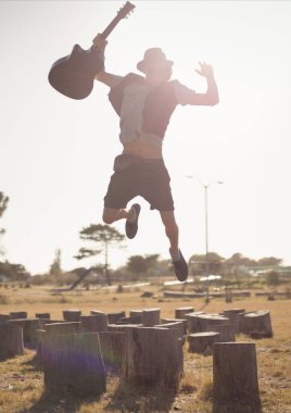 Güneşli bir günde elinde gitar tutan bir adam ağaç kütüklerinde zıplıyor.