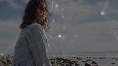Kayalık sahilde duran, yüzünü eğen, esintiyi hisseden, ışıldayan teknoloji ağını harekete geçiren bir kadın. Deniz manzarası, bağlantı, teknoloji, dinginlik, soyutlama, keşif, ufuk.