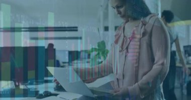 Dizüstü bilgisayar ekranına bakmak için öne eğilen kadın, basılı iş raporunu grafiklerle örtüyor. Profesyonel, üretkenlik, minimalist, veri, işbirliği, çalışma alanı, dijital