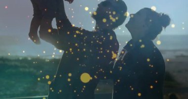 Ebeveynler gün batımında deniz kenarındaki korkuluklarda altın parıltılara sebep olan pazarlama için bebek kaldırıyorlar. Aile, ebeveynlik, kutlama, neşe, birliktelik, deniz kenarı, sükunet