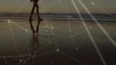 Islak kıyı şeridine adım atan ve yürüyen bir kadın, gün batımına karşı örgü ören ağ teknolojisini gösteriyor. Doğa, dinginlik, dijital, geleceksel, yansıma, sükunet, macera