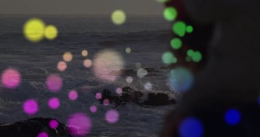 Orta yaşlı bir kadın akıllı telefonuyla kayıt yapıyor. Işık küreleri dalgaların üzerinde süzülürken. Deniz manzarası, sükunet, alacakaranlık, ortam, minimalizm, tefekkür, doğa