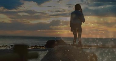 Kadın gün batımında kıyıdan kayaya adım atıyor, çağrıyı bitiriyor, sağlık parıltılarıyla tansiyonu düşürüyor. Manzaralı, düşünceli, sakin, siluet, doğa, rahatlama, farkındalık.