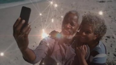 Üst düzey bir çift sahilde selfie çekmek için ağ üzerinden akıllı telefon kaldırıyor. Uygulamada galeri inceleniyor. Bağlantı, nostalji, teknoloji, samimiyet, rahatlama, arkadaşlık, sıcaklık.