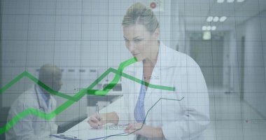 Writing senior doctor wearing white lab coat in hospital hallway, with clipboard and green chart. Medical, healthcare, professionalism, collaboration, clinical, diagnostics, documentation