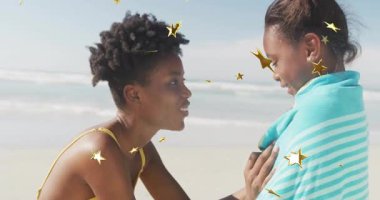 Anne turkuaz havlu tutuyor ve çocuğu sahilde kurutuyor. Sağlık gösterisinde altın yıldızlar sürükleniyor. Aile, beslenme, plaj, yaz, su, sıcaklık, rahatlama.