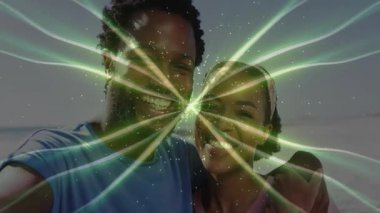 Kıyıda akıllı telefon tutan çift selfie çekiyor ve yeşil fraktal ışınlar teknoloji için yanıp sönüyor. Katı, dijital, canlı, çağdaş, enerjik, sembolik, bağlantı