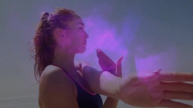 Plajda kolunu uzatan bir kadın pembe-mor enerji puslu ön koluyla fitness demosunu başlatıyor. Atletik, farkındalık, canlılık, enerji, dinginlik, modern, dinamik