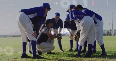 Huddling softball team listening to coach kneeling with clipboard in outfield, blue jerseys, gloves. Team, coaching, athletic, outdoor, training, teamwork, vibrant