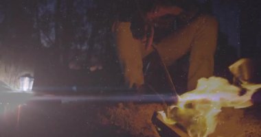 Alevli sopayla ateş közlerini iten adam kamp pişirmek için ızgara kavurucu çubuğu hareket ettiriyor. Açık hava, kamp, vahşi doğa, akşam, köy, doğa, macera.