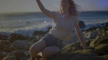 Sosyal medya kullanıcısı diz çöküp akıllı telefon selfie 'sini çekiyor ve dalgalar üzerinde hareketli grafikleri aktifleştiriyor. Sahil, sahil, teknoloji, dijital sanat, yaşam tarzı, gün batımı, rahatlama