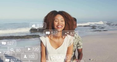 Kumsalda gülümseyen bir kadın sosyal medya ikonlarını aktifleştiriyor ve kucaklaşan bir çiftin etrafındaki nişanı yansıtıyor. Romantik, bağlantı, açık hava, canlı, mutluluk, sevgi, kutlama