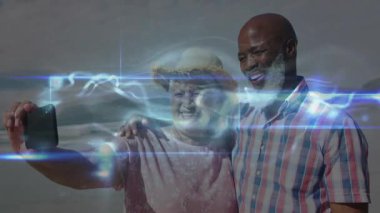 Kıdemli çift selfie çekmek için telefonları kaldırırken ışık çizgileri sahilde teknolojiyi gösteriyor. Romantizm, bağlantı, dinginlik, dijital alarm, yaşam tarzı, açık hava, uyum.