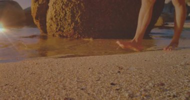 Kum tepesinde yükselen bacakları gösteren bir seyahat klibi. Güneşli kayaları aşıp sığ dalgalara doğru yürüyor. Kıyı, yalınayak, gün batımı, kıyı şeridi, dalgalar, ayak izleri, boş zaman